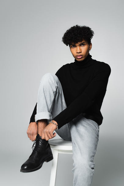 trendy african american man in black turtleneck and jeans sitting on white stool and looking at camera on grey background - Foto, Imagen