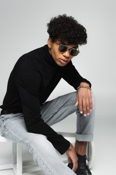african american man in jeans, black turtleneck and dark sunglasses sitting on overturned stool on grey  - Foto, immagini