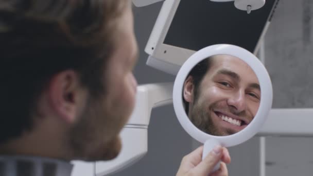 Boldog férfi beteg mosolyog tükörreflexió, élvezi az eredményt a fogfehérítés eljárás fogászati klinikán, közelről - Felvétel, videó