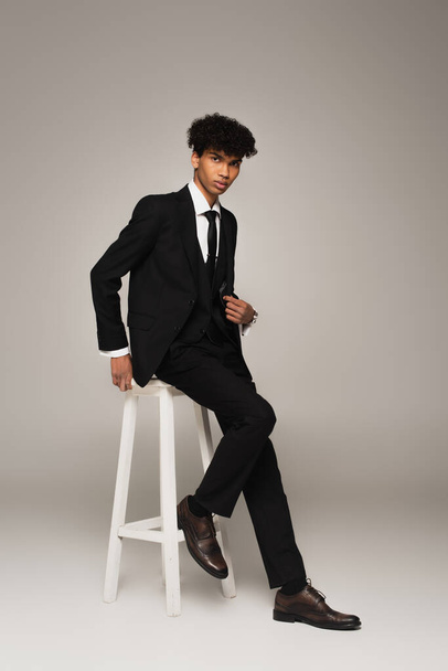 full length view of african american man in elegant formal wear sitting on high stool on grey background - 写真・画像
