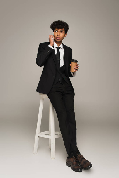 african american businessman sitting on high stool with coffee to go and talking on cellphone on grey background - Photo, Image