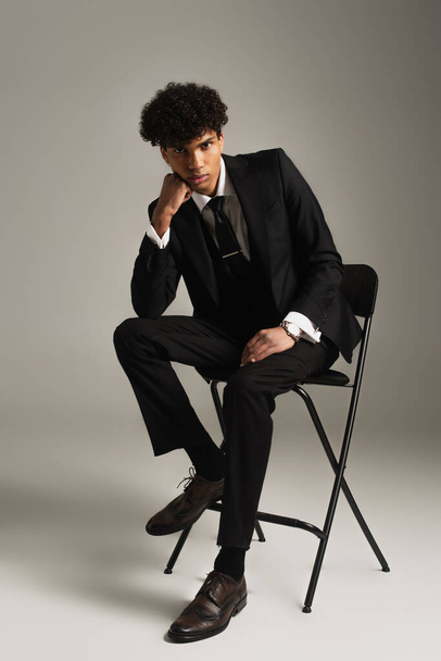 modny Afroamerykanin mężczyzna w eleganckim garniturze siedzi na krześle z ręką blisko twarzy na szarym tle - Zdjęcie, obraz