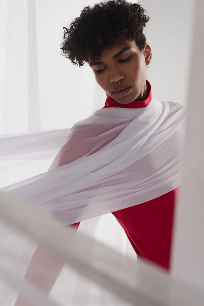 stylish african american man wearing red turtleneck posing in white tulle drapery - Photo, Image