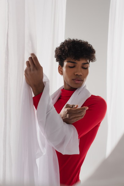 curly african american man in red turtleneck posing near white chiffon cloth - Foto, Imagen