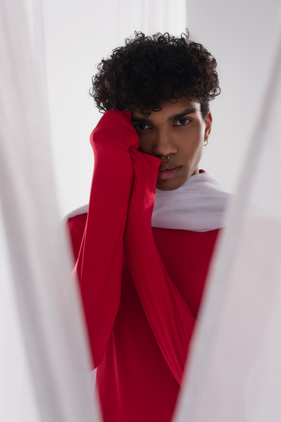young african american man in red turtleneck posing near white chiffon cloth with hands near face - Foto, Bild