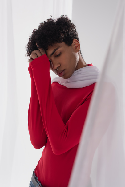 african american man in red turtleneck posing near white tulle cloth with closed eyes and hands near face  - 写真・画像