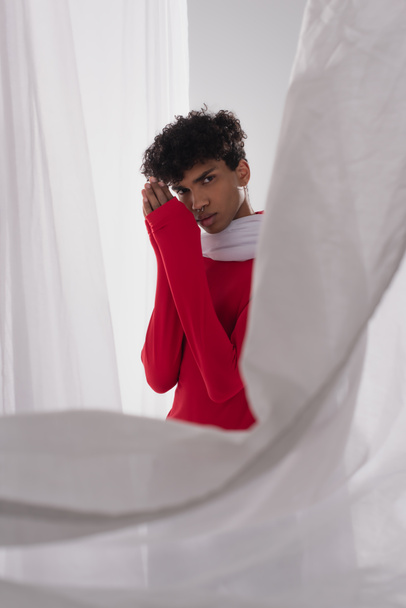 stylish african american man in red turtleneck posing with praying hands near white blurred drapery - Foto, Imagem