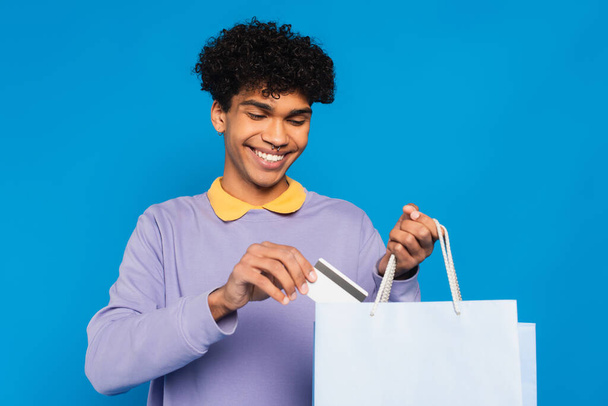 joyful african american man putting credit card into shopping bag isolated on blue - Foto, afbeelding