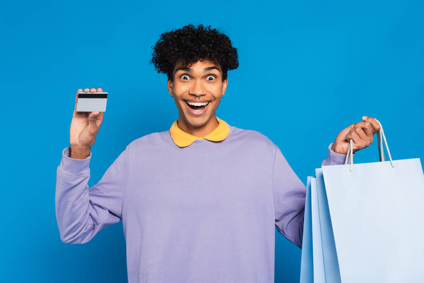 amazed african american man holding shopping bags and credit card isolated on blue - Foto, Imagen