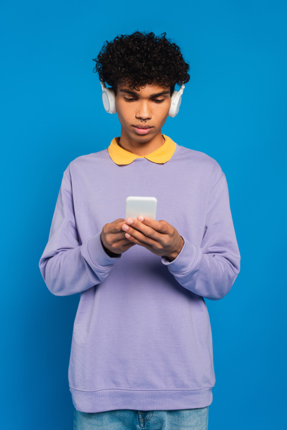 young african american man in headphones using cellphone isolated on blue - Fotoğraf, Görsel