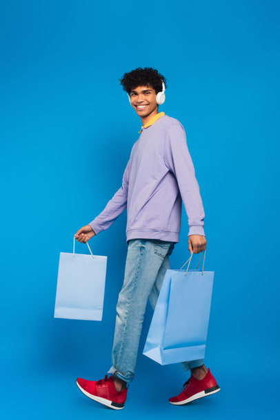 smiling african american man in headphones walking with shopping bags on blue background - Fotografie, Obrázek