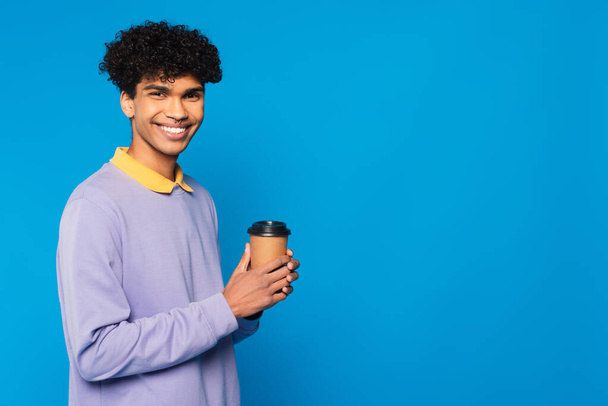 cheerful african american man in purple jumper with yellow collar holding paper cup isolated on blue - Zdjęcie, obraz