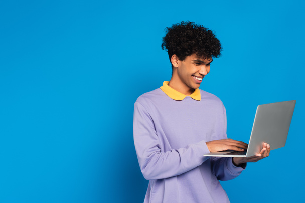 happy african american man in violet pullover using laptop isolated on blue - Foto, Bild