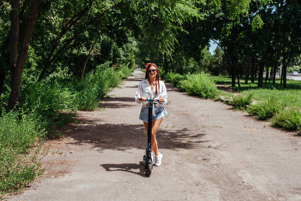 Χαριτωμένη μελαχρινή κοπέλα βόλτες σε ένα σκούτερ ηλεκτρόδιο στο πάρκο φοράει ένα λευκό πουκάμισο. έννοια των οικολογικών μεταφορών και ενοικίου.  - Φωτογραφία, εικόνα
