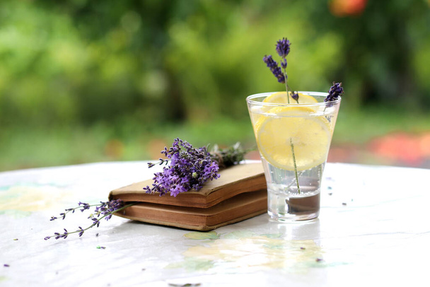 Homemade lemonade with lemon and lavender in a glass glass on a table in the garden with a niga and a bouquet of lavender. Cooling tonic soothing detox. - Photo, image