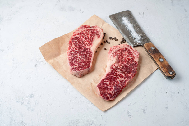 Two raw fat and juicy beef steaks on craft paper with pepper and a meat cleaver - 写真・画像