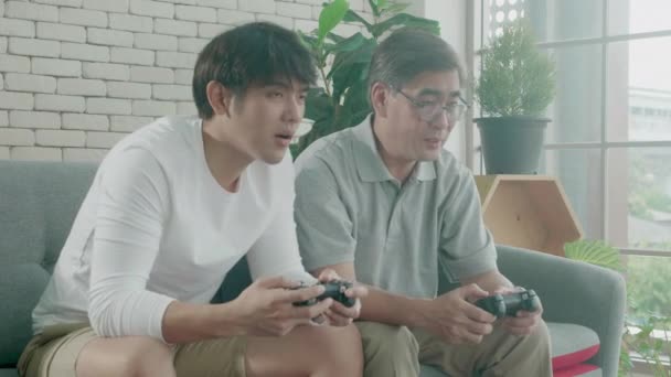 Aasialainen perhe isä ja poika pelata videopeli sohvalla kotona, vanhukset ja nuori mies kahden sukupolven kilpailu nauttia joystick viihdettä ja innoissaan, elämäntavat suhde käsite. - Materiaali, video