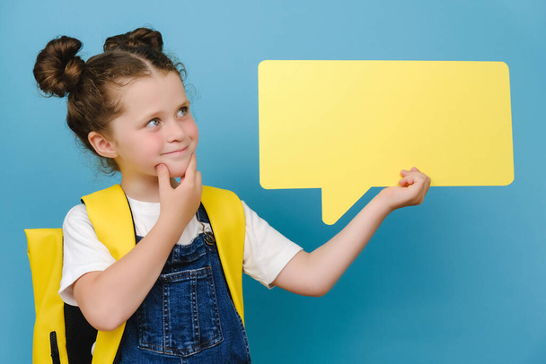 Portrait of thoughtful smiling little caucasian schoolgirl holding yellow blank speech bubble above head, wears backpack, posing isolated over blue studio background with copy space for advertising - Φωτογραφία, εικόνα