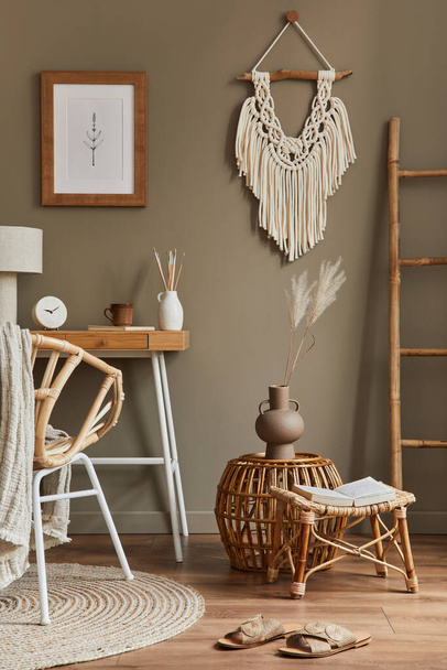 Stylish bohemian interior of home office space with wooden desk, rattan armchair, brown mock up poster frame, macrame, office supplies, lamp, decoration and elegant personal accessories in home decor. - Foto, Imagem