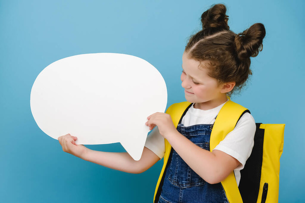 Portrait of interested cheerful kid hold white paper blank speech bubble, thoughts about future weekends, wear stylish yellow backpack, isolated over blue color background in studio. Back to school - 写真・画像