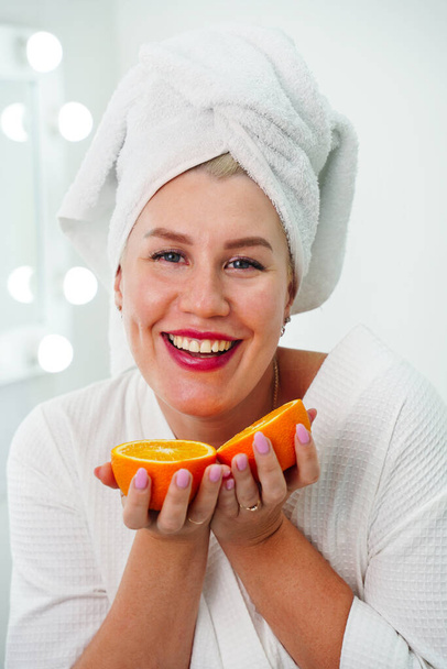 Mladá šťastná energická žena v bílém rouchu a ručník na hlavě drží v rukou polovinu pomerančů v blízkosti očí v koupelně, vitamín C v kosmetice - Fotografie, Obrázek