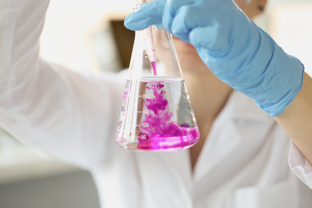 Химик держит фляжку с прозрачной жидкостью в руках и капает в нее фиолетовый растворитель - Фото, изображение