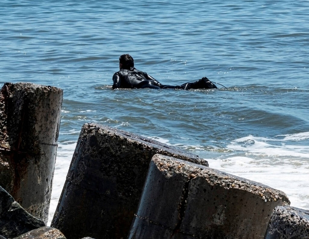 Um homem está deitado em sua prancha de surf ao lado de um molhe vestindo um terno preto esperando por uma cera para montar - Foto, Imagem
