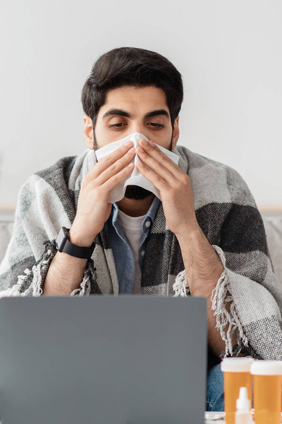 Travail à distance. Malade gars arabe ayant rhinite éternuer et le nez qui coule, travaillant sur ordinateur portable à la maison, culture - Photo, image