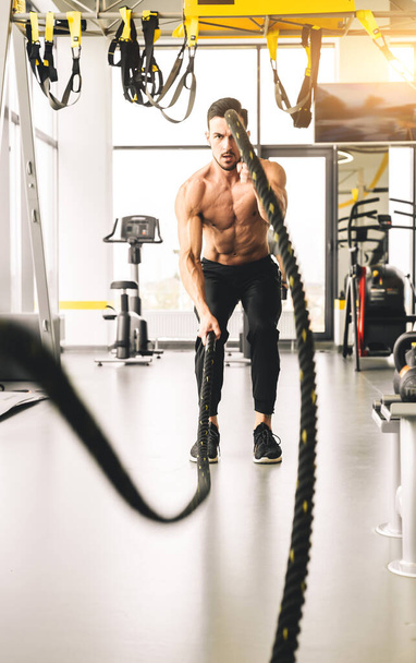 Handsome young sporty male bodybuilder with naked torso doing exercises with ropes in the gym - Foto, Imagem