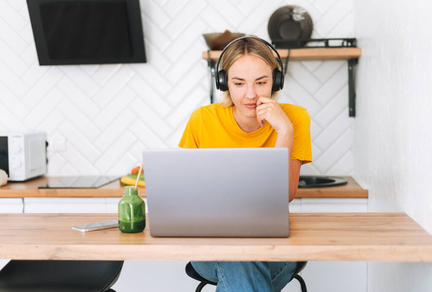 Young smiling blonde woman in yellow t-shirt with headphones working at laptop in kitchen at home - Photo, Image