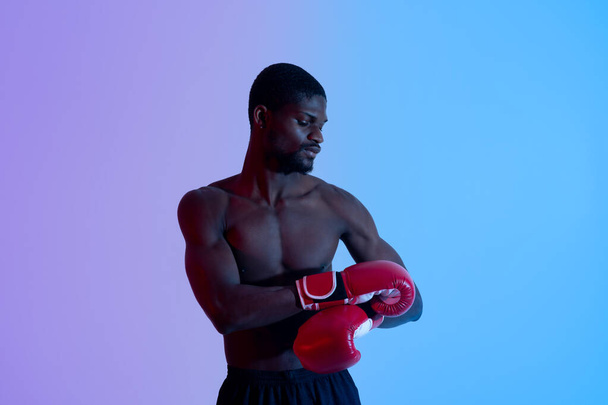Professionele jonge zwarte bokser met kale romp het aantrekken of uittrekken van rode handschoenen in neon verlichting - Foto, afbeelding