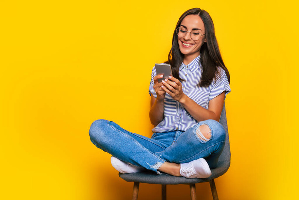 Happy beautiful stylish woman is sitting on the chair and using her smart phone while she is isolated on yellow background - Foto, imagen