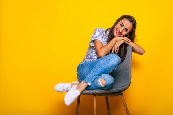 Cute smiling stylish woman is sitting on the chair and posing isolated on yellow background - Foto, imagen