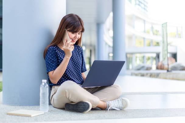Happy young Asian woman sitting and talking on her phone while using her laptop computer outside of her office building, modern lifestyle concept - Фото, изображение