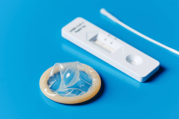 um preservativo enrolado e um dispositivo de teste de diagnóstico do antigénio covid-19, com um resultado negativo, num fundo azul - Foto, Imagem