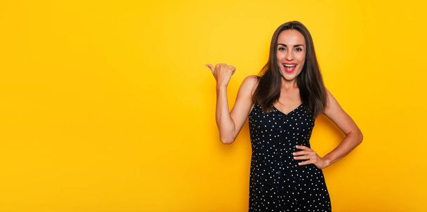Happy excited gorgeous smiling brunette woman in a black summer dress is posing isolated on yellow background and pointing away - Valokuva, kuva