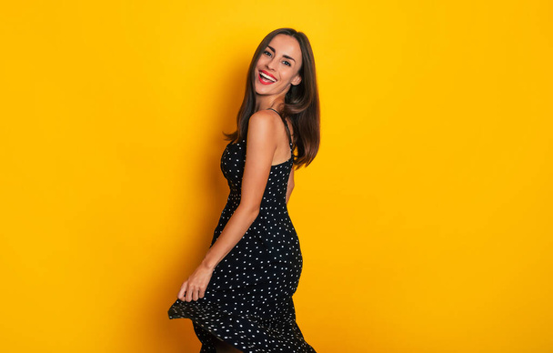 Happy excited gorgeous smiling brunette woman in a black summer dress is dancing isolated on yellow background and having fun - Valokuva, kuva