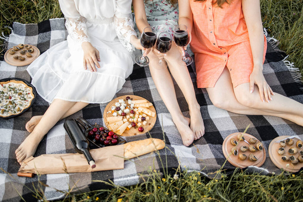 Three girls, sitting on a blanket on nature, having picnic in a city park with a glasses of wine - 写真・画像