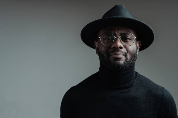 Handsome african american man in a hat wearing glasses and in a black turtleneck smiling looking at the camera - Zdjęcie, obraz