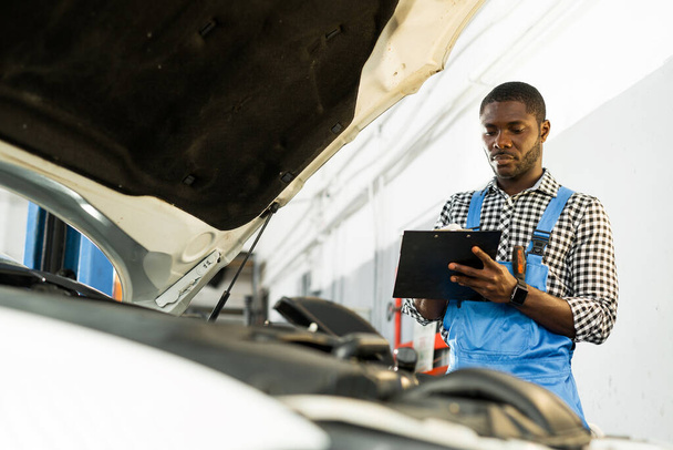 Serious african american mechanic holds a notebook in his hand while standing near the open hood of a car writing down problems - Foto, Imagem
