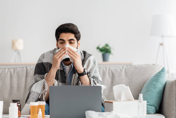 Beteg arab férfi tüsszentés orr papírzsebkendő és nézi laptop képernyőn, ül otthon és dolgozik, üres hely - Fotó, kép