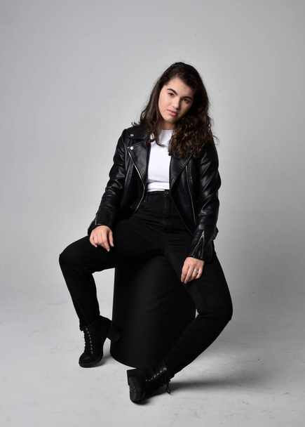 Full length portrait of young woman with natural brown hair,  wearing black leather scifi outfit with jacket, sitting pose on light studio background. - Foto, Imagen