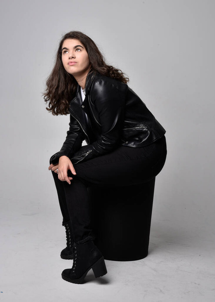 Full length portrait of young woman with natural brown hair,  wearing black leather scifi outfit with jacket, sitting pose on light studio background. - Fotoğraf, Görsel