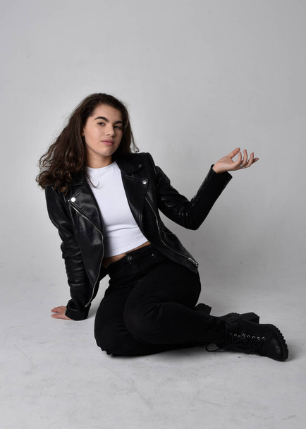 Full length portrait of young woman with natural brown hair,  wearing black leather scifi outfit with jacket, sitting pose on light studio background. - Фото, зображення