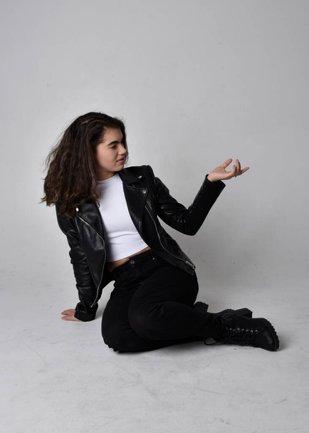 Portrait pleine longueur de jeune femme aux cheveux bruns naturels, portant une tenue scifi en cuir noir avec veste, posant assise sur un fond studio léger. - Photo, image