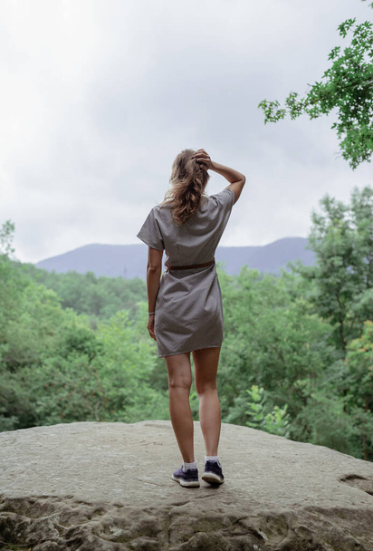 Concepto de viaje y libertad. Vista trasera de una mujer joven en vestido de verano de pie sobre una gran roca en el bosque mirando hacia otro lado - Foto, imagen