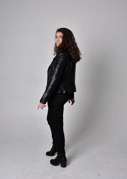 Портрет молодой женщины с натуральными каштановыми волосами, в черной кожаной одежде с курткой, стоящей в позе спиной к камере, на светлом студийном фоне. - Фото, изображение