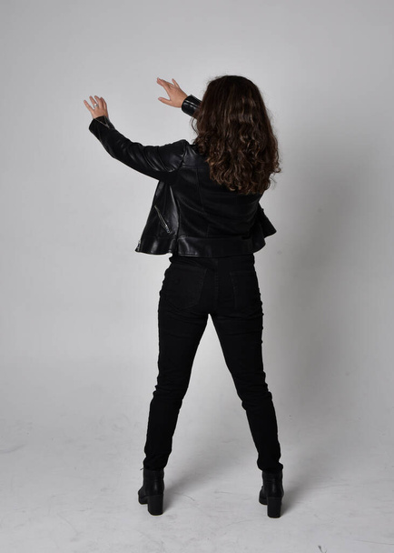 Full length portrait of young woman with natural brown hair,  wearing black leather scifi outfit with jacket, standing pose  with back to the camera, on light studio background. - Foto, Imagem