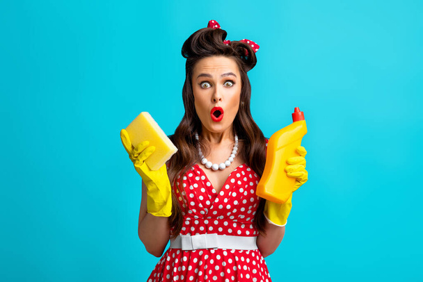 Schockierte Pinup-Frau in gepunktetem Kleid mit Toilettenwaschmittel und Schwamm, macht überraschtes Gesicht auf blauem Hintergrund - Foto, Bild