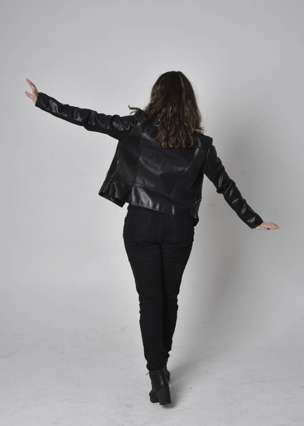 Full length portrait of young woman with natural brown hair,  wearing black leather scifi outfit with jacket, standing pose  with back to the camera, on light studio background. - Фото, зображення